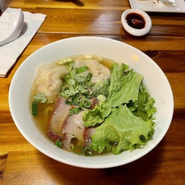 A bowl of Vietnamese wonton noodle soup with sliced pork, lettuce, and herbs on a wooden table.