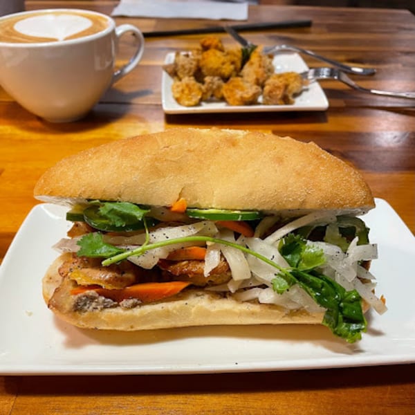 Vietnamese bánh mì sandwich with a side of fried chicken and a latte on a wooden table.